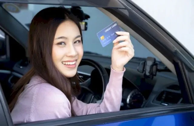 Счастливая красивая азиатская женщина сидит внутри нового автомобиля синего цвета и показывает кредитную карту